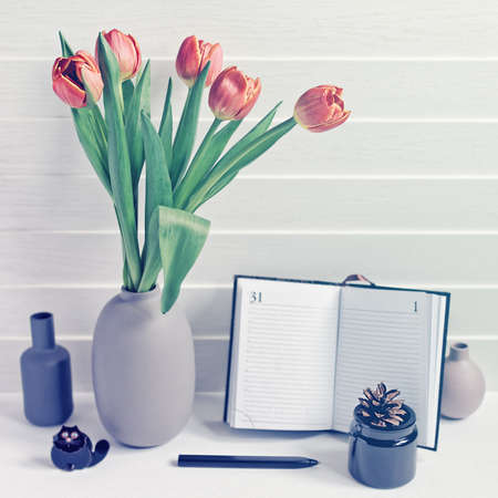 An open notepad with a bookmark, a black pen, a pine cone in a glass container, a cat figure and a bouquet of red tulips in a vase are on a wooden background.Image suitable for teacher's dayの写真素材