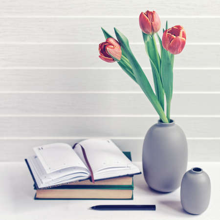 A bouquet of three red tulips is in a gray vase. It's a stack of books, an open notebook and a black pen. White wooden background. The image is suitable for a writer, teacherの写真素材