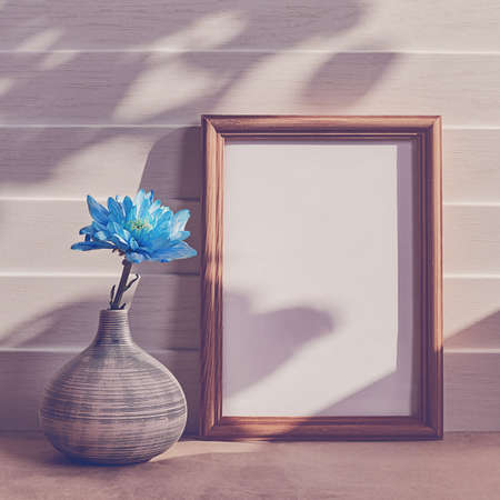A flower of a blue chrysanthemum or chamomile is in a striped vase. Behind there is a large wooden frame. White wooden background.の写真素材