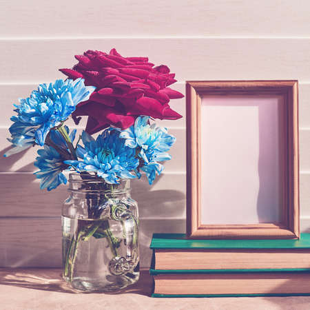 A red fresh rose along with blue chrysanthemums or chamomiles is in a glass jar. There is an empty...