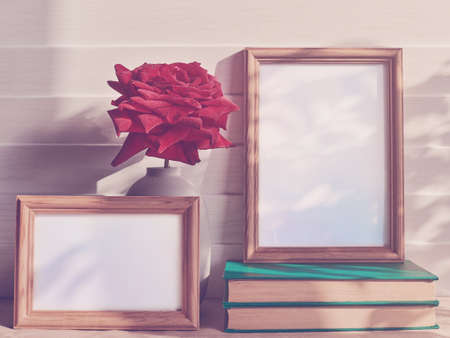 A fresh red rose with droplets on the petals is in a gray opaque vase. There are two empty wooden frames, one of which is on the pile of books. White wooden backgroundの写真素材