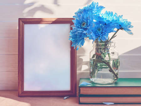 An empty wooden frame. The distance is a glass jar with blue chrysanthemums or daisies on a stack of books.の写真素材