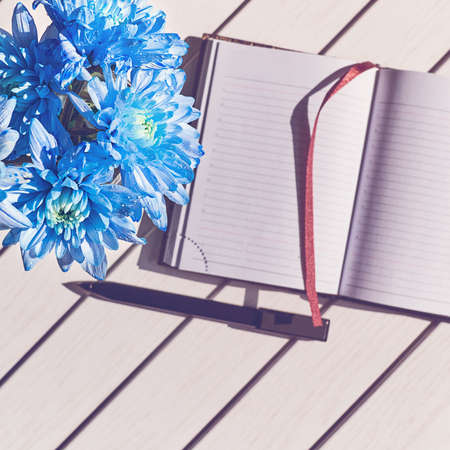 Open notepad with fabric bookmark, black pen and bouquet of blue chrysanthemums are on a white...