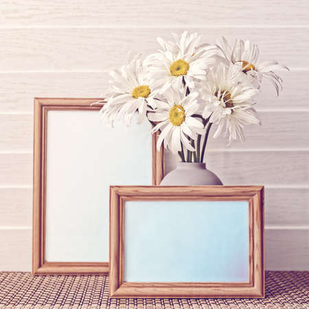 White chamomiles are in a gray vase. There is an empty wooden frame. White wooden background.の写真素材
