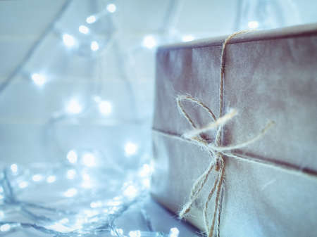 A large gift box is wrapped in paper and bandaged with string. On a wooden background there is a flickering garland.の写真素材
