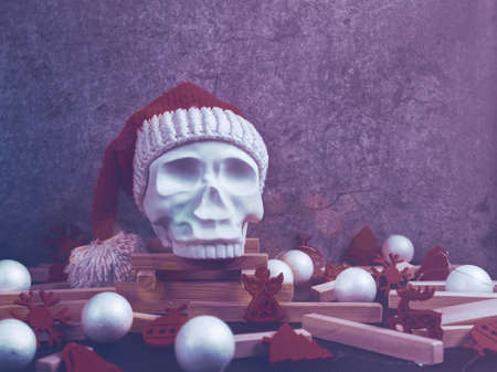 A red Santa Claus hat is put on a decorative sculpture in the form of a human skull. Nearby are balls and figures in the form of a Christmas tree. Gray cement background with cracks and scuffs.の写真素材