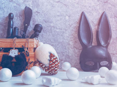 Various sex toys are in a wooden box. Nearby is a large Christmas fir cone and a black mask. Balls are scattered nearby. Gray background with cracks and scuffs. Minimal composition.の写真素材