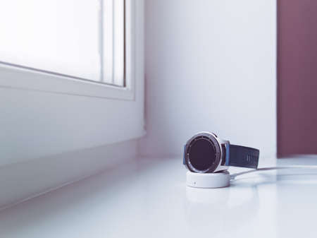 Wireless charging gadgets. A smart watch with a blue silicone strap is located in the charging cradle on the windowsill.の写真素材