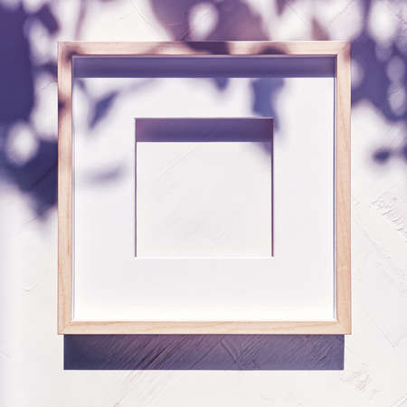 An empty wooden photo frame with a passepartout is on a white textured background with shadows from the foliage. Flat lay. Copy space.の写真素材