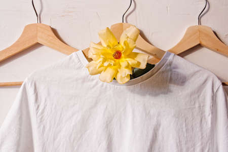 Creative floral background. A white cotton T-shirt is hung on a wooden coat hanger and decorated with a yellow flower. Fashionable layout.の写真素材
