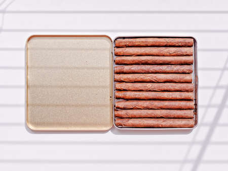 Cigarillos are in an open tin box. A shadow from the blinds falls on a white background. Tobacco...