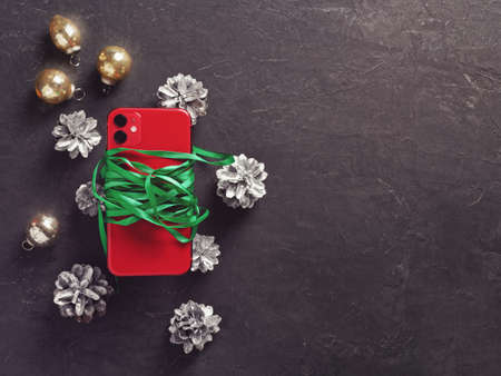 A red smartphone with two cameras is wrapped in a green ribbon and is located on a dark cement...