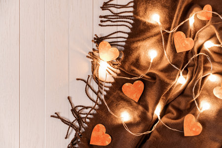 Paper hearts and a glowing garland are on a brown fringed scarf on a light wooden background. Composition for Valentine's Day. Top view. Flat lay. Copy space.の写真素材