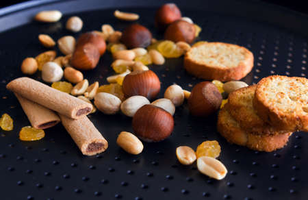 Peanuts, hazelnuts, pistachios, yellow raisin, jam sticks, baguettes on a black baking pan with holes. Selective focusの写真素材