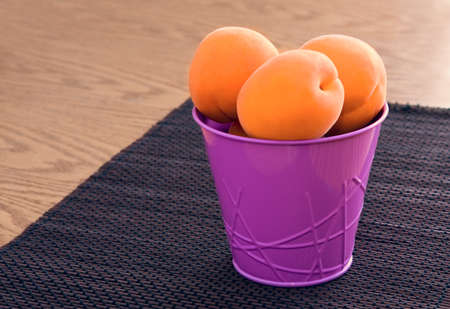 Sweet ripe yellow apricots in a small metallic violet pink bucket on a small bamboo mat on a wooden background. Selective focus.の写真素材