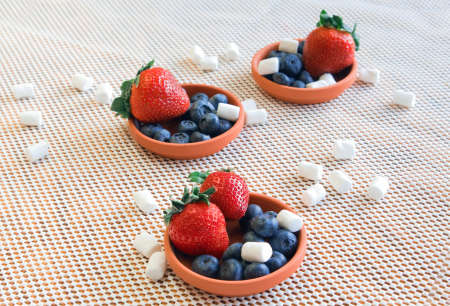 Red strawberries with green leaves, blueberries and white sweet marshmallows in small clay dishes on a white background. Selective focus.の写真素材
