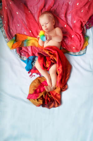 Magical cute blond baby girl lying on a blue bed sheet covered with beautiful red, yellow, orange...