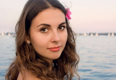 Closeup portrait of a young girl with a pink flower in hair on a beach near the waterの写真素材