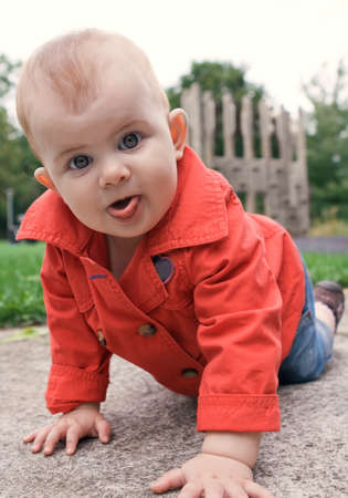Portrait of a cute baby in red trench and jeans showing her tongue crawling on a playground outdoorの写真素材