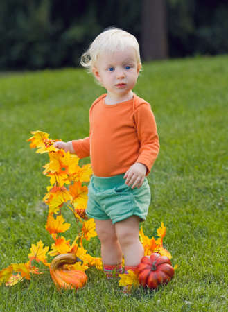 Portrait of a cute funny adorable blond Caucasian baby toddler with blue eyes in orange shirt and green shorts standing in the grass on the field meadow with yellow autumn fall leaves with pumpkins  Halloween cardの写真素材