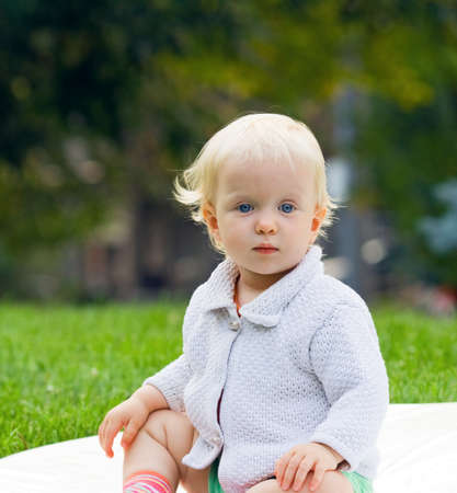 Closeup portrait of a cute adorable child baby with blond hair and blue eyes, caucasian, in a knitted wool jacket sitting on a blurry background with green leaves, outdoor  Summer, autumn, copy space for textの写真素材