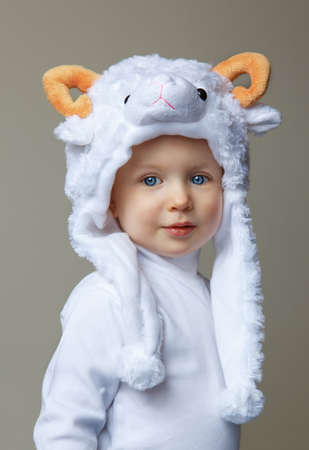 Cute adorable pretty Caucasian baby toddler with large blue eyes wearing a sheep hat hood with yellow horns on top and white shirt standing on a light background looking into the camera, New Year 2015 concept, studioの写真素材