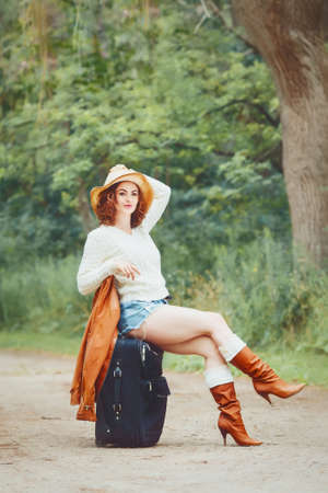 Portrait of a beautiful Caucasian young girl woman in white sweater, high leather brown boots, blue jeans shorts and straw hat, sitting on travel back on the country road, travel wonderlust adventure vacation conceptの写真素材