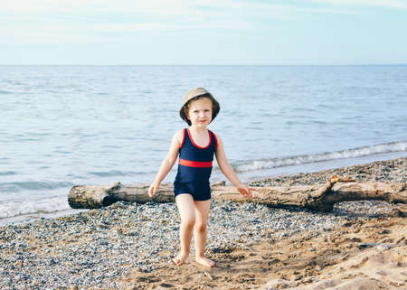Portrait of adorable white Caucasian one toddler little girl with hat on her head going on lake sea gravel beach, emotional face expression, lifestyle summer moodの写真素材