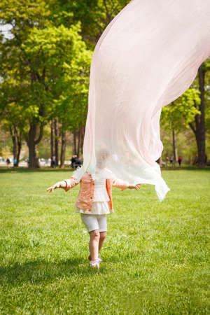 Portrait of a cute adorable baby toddler girl playing on field meadow on bright sunny summer day with mom who covers her with transparent light cloth scarf making her look like a ghost, happy childhood family activity lifestyle conceptの写真素材