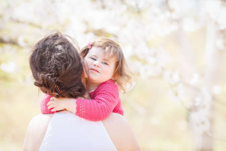 Close up portrait of caucasian mother in white dress holding hugging her daughter in pink clothes with funny face expression on sunny spring summer day in the park outsideの写真素材