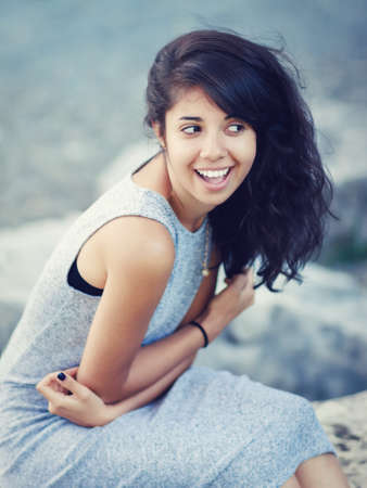 Portrait of beautiful Hispanic latino white girl woman with brown eyes, long dark curly wavy hair in gray dress sitting in park outside smiling laughing looking away, lifestyle portrait conceptの写真素材