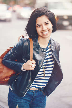 Portrait of beautiful smiling young Caucasian latin girl woman with dark brown eyes, short dark hair, in blue jeans, leather biker jacket with backpack in city outsideの写真素材