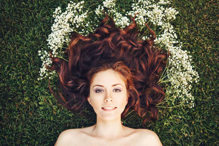 Close up portrait of young beautiful girl woman with red brown hair laying on grass with white small flowers around her head .View from above top overhead.の写真素材