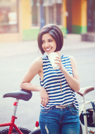 Portrait of beautiful smiling young hipster latin girl woman with short hair bob, in blue jeans, striped tshirt, holding cup of coffee wth bike in city looking awayの写真素材