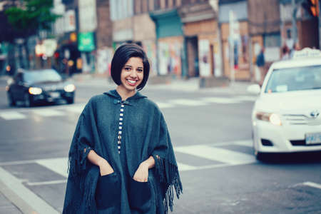 Portrait of beautiful smiling young hipster latin hispanic girl woman with short hair bob, in torn jeans, grey blue poncho cape, standing outside in city streetの写真素材