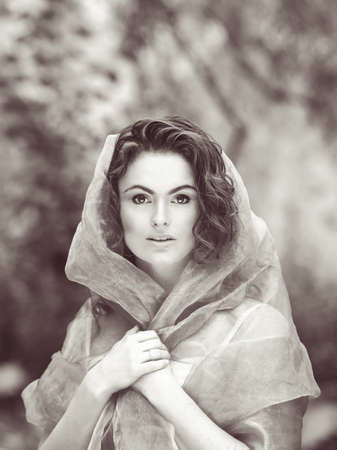 Black and white close-up beauty portrait of young white Caucasian girl with wavy curly hair covered with hood clothes gauze, in park outside on summer dayの写真素材