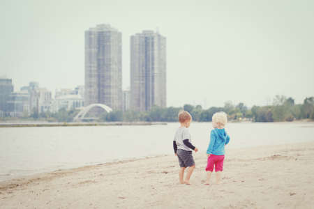 Two children kids toddlers boy girl walking on sand sea lake ocean beach looking for something interesting,  picking seashells, talking and playing together, best friends forever, toned with  filters, retro old vintage effect, hipster style, everyday lifeの写真素材
