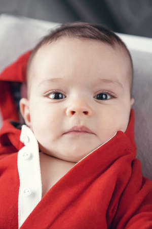 Closeup portrait of cute adorable Caucasian baby boy girl with black brown eyes in red hoodie shirt on changing table looking directly in camera, natural light indoorsの写真素材