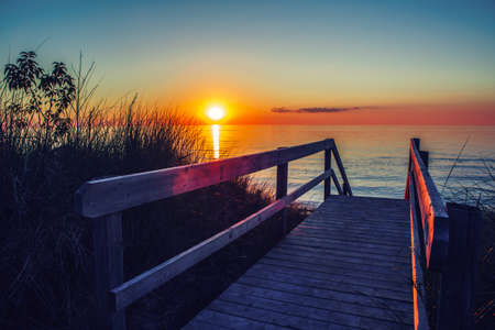 Beautiful evening sunset landscape at Canadian Ontario lake Huron in Pinery Park, orange blue red sky sun. Amazing summer sunset view on the beach. Dunes, grass, wooden stair leading to waterの写真素材