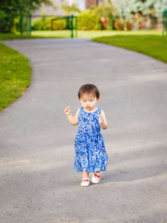 Portrait of a cute adorable little Asian girl child, one two years old, in blue dress and white shoes walking running in park outside on summer day, concept of first baby stepsの写真素材