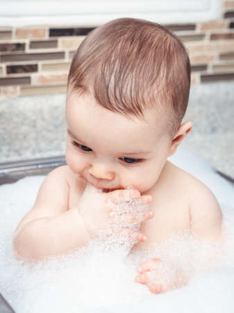 Portrait of cute Caucasian funny baby girl boy with dark black eyes sitting in big kitchen sink with water and foam near window looking away, lifestyle everyday conceptの写真素材