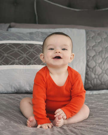 Portrait of cute adorable Caucasian smiling laughing baby boy girl with black brown eyes in orange red onesie shirt sitting on bed looking directly in camera, natural window light, lifestyleの写真素材