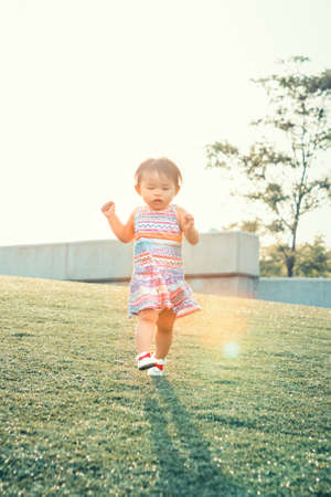 Portrait of a cute adorable little chinese asian vietnamese girl child, one two years old, in dress with ornaments and white shoes, running on field meadow grass backlit with sunset, concept of childhood lifestyleの写真素材