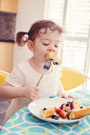 Portrait of one happy white Caucasian kid girl toddler with pig-tails in white dress eating breakfast waffles fruits with fork in sunny kitchen early morningの写真素材