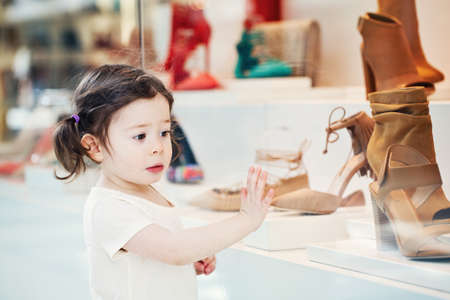 Closeup portrait of cute adorable sad upset white Caucasian toddler girl child with dark brown eyes and curly pig-tails hair in white light dress tshirt in mall looking at shoes in shop store windowの写真素材