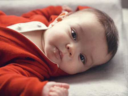 Closeup portrait of cute adorable Caucasian baby boy girl with black brown eyes in red hoodie shirt on changing table looking directly in camera, natural light indoorsの写真素材