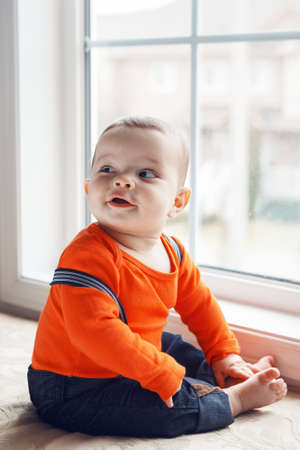 Portrait of cute adorable Caucasian baby boy with black eyes in orange shirt  , jeans with suspenders barefoot sitting on windowsill looking away, natural window light, lifestyleの写真素材