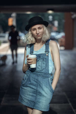 Portrait of beautiful Caucasian teenage young blonde alternative model girl woman in blue tshirt, jeans romper looking in camera holding cup of coffee, toned with filtersの写真素材
