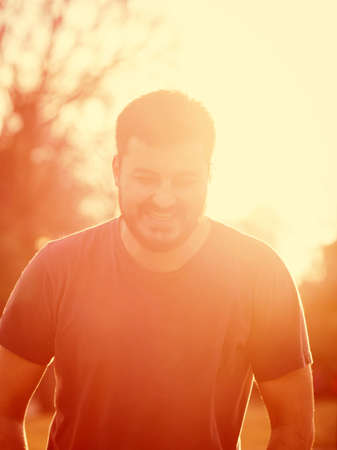Portrait of a young bearded man in t-shirt laughing smiling at sunset light, blurry background, toned with filtersの写真素材