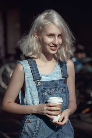 Portrait of beautiful Caucasian teenage young blonde alternative model girl woman in blue t-shirt, jeans romper looking in camera holding cup of coffee, toned with filtersの写真素材